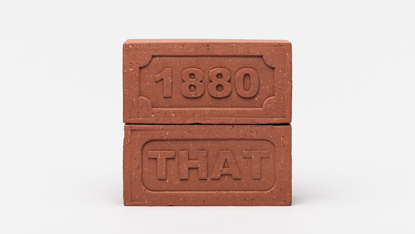 A sculpture that consists of two brown rectangular building bricks stacked on top of each other. The top brick is embossed with the number 1880 and the bottom brick is embossed with the word THAT.