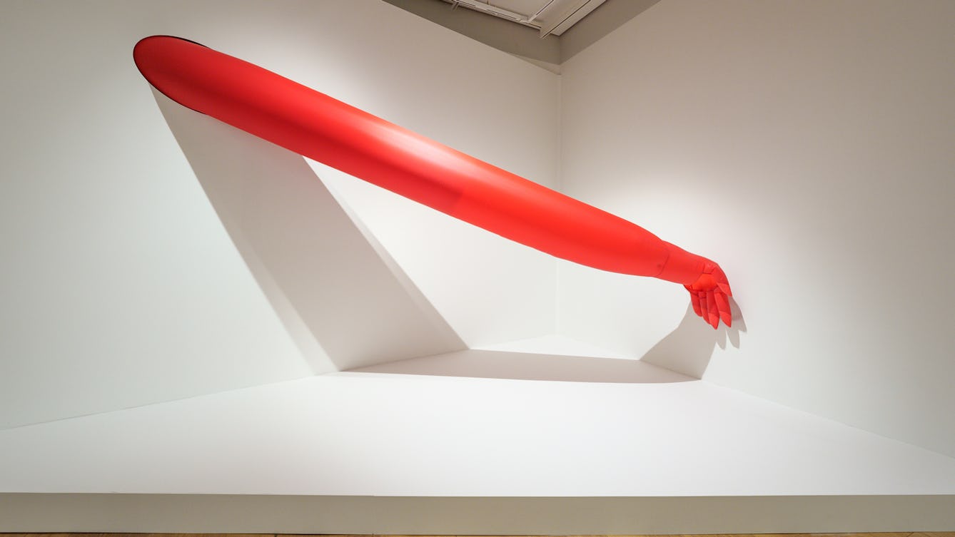 A large inflatable arm emerging from a hole in the wall in a corner of an exhibition gallery. The arm is fully stretched out and the back of the hand is resting on the adjacent wall.