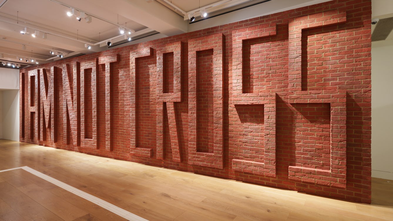 A red brick wall constructed inside an exhibition gallery. The wall has raised bricks across the surface, spelling out the phrase "I AM NOT CROSS".