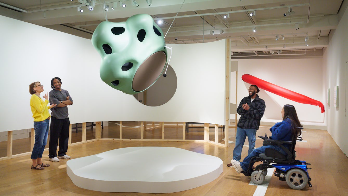Four people stand around a suspended sculpture in the exhibition '1880 THAT' in pairs, talking about the work and looking at it.