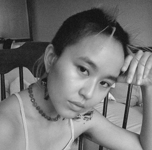 A black and white photograph of the head and shoulders of a young East Asian person. They are leaning their head on one hand and looking sideways into the camera.