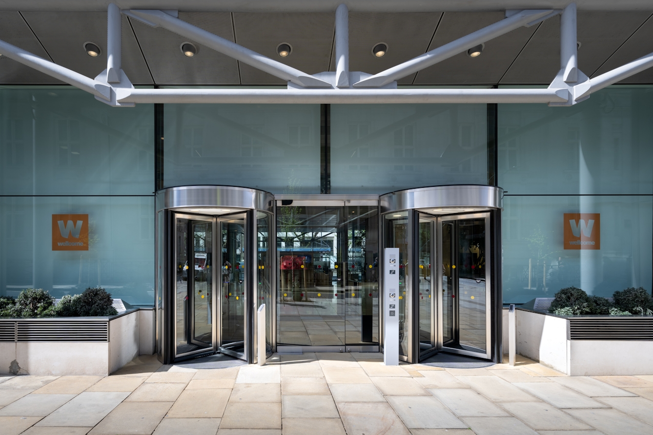 Three large doors in a row. The two outer doors are glass revolving doors, each with the sign 'wellcome collection' next to them. The central doorway is wider and has a plain glass door.
