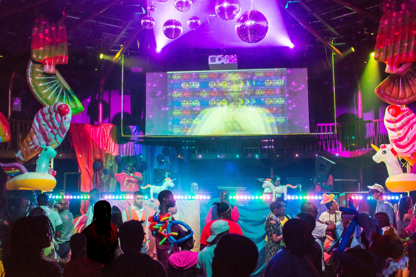 Photograph of the Beautiful Octopus Club. At the club night people are wearing vibrant, colourful clothes, there are bright lights, balloons, inflatable unicorns, disco balls and DJs on stage. 