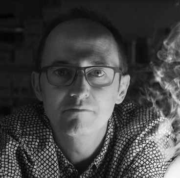 Black and white photograph of the head and shoulders of a middle-aged man with glasses. He is looking directly at the camera.