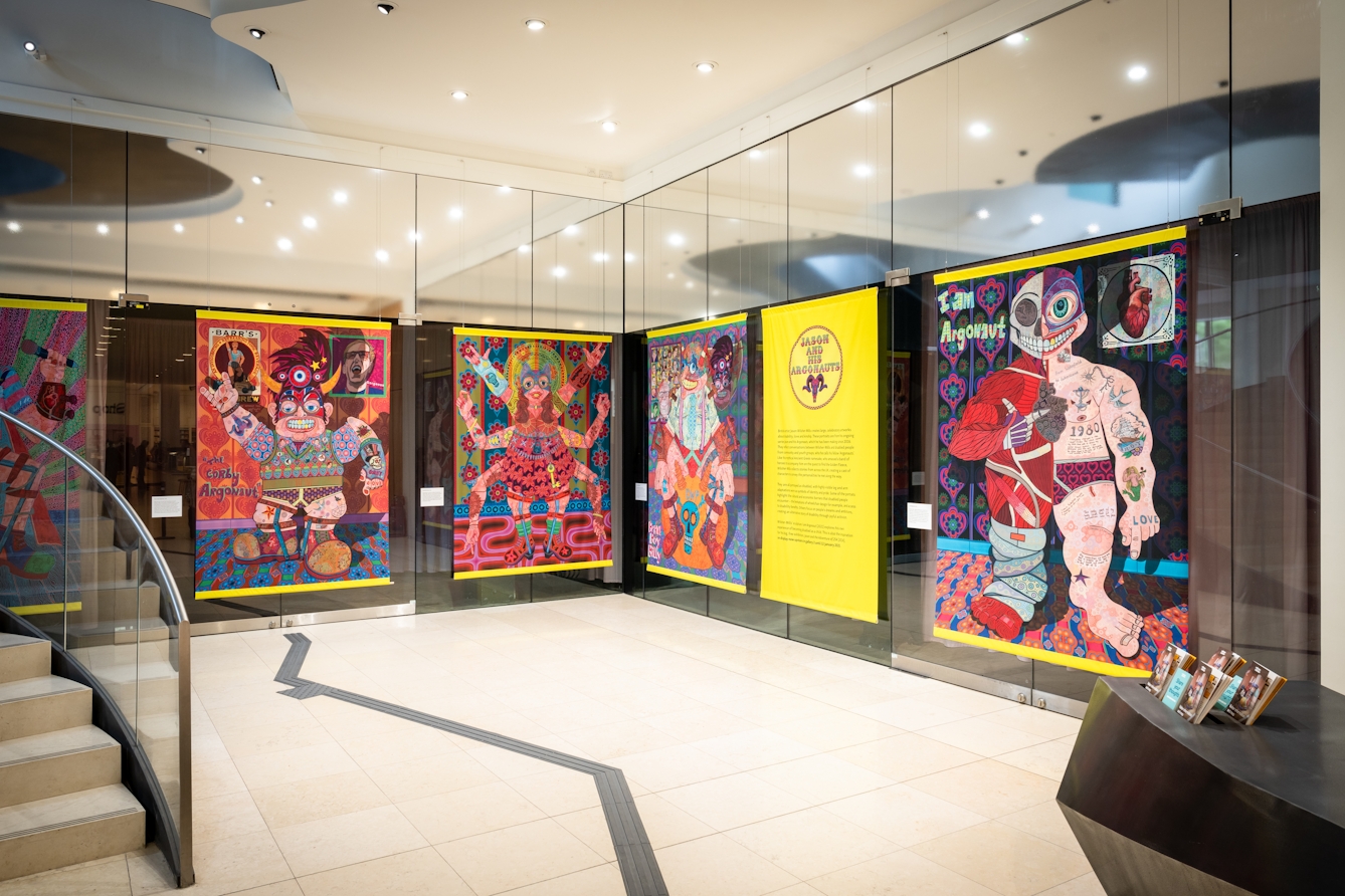 A series of large-scale, colourful artworks by Jason Wilsher-Mills on display in the Atrium at Wellcome Collection. Part of a staircase can be seen on the left and part of a welcome desk on the right. There is a thick, tactile black line on the floor, which can be followed to reach each of the artworks.