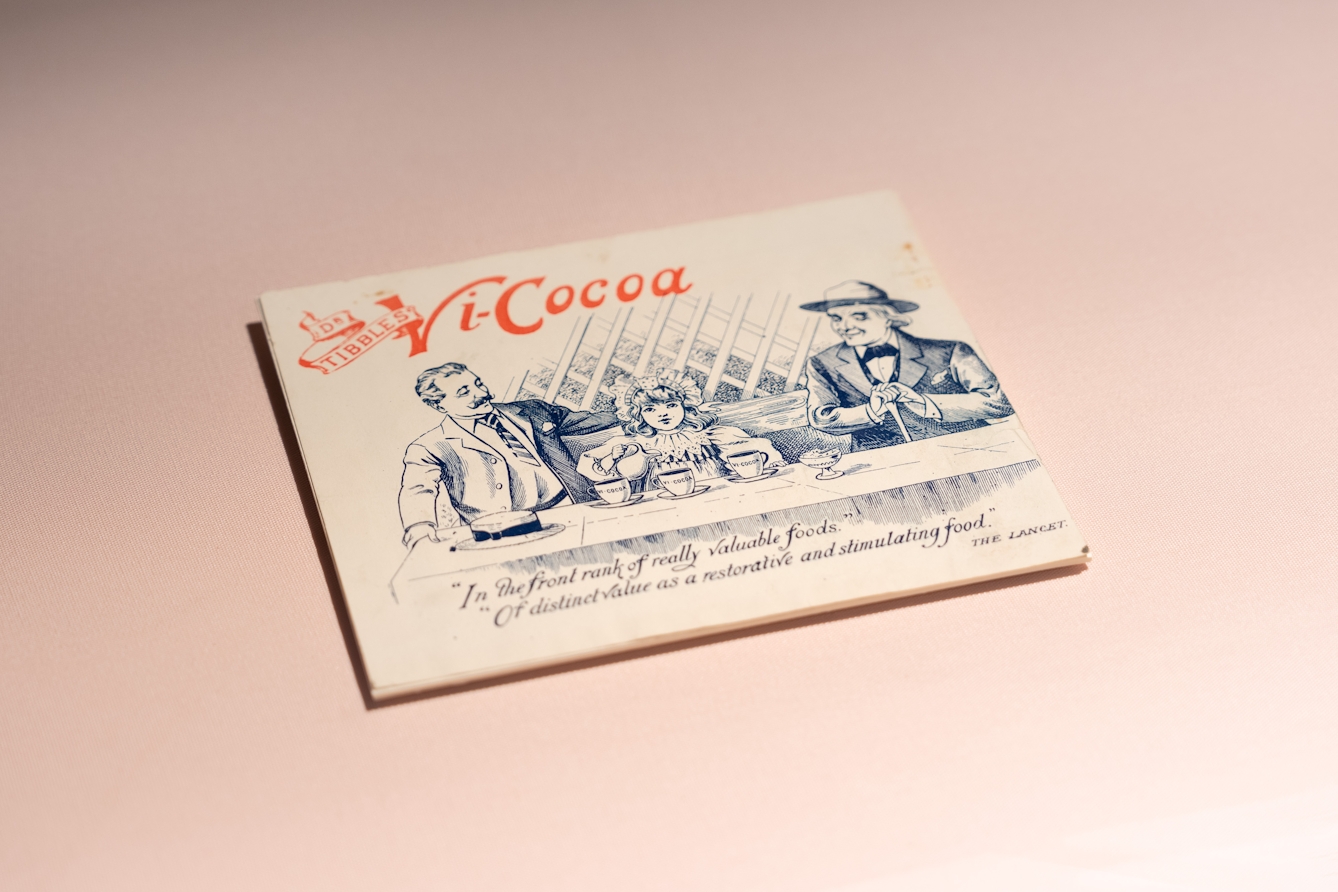 Photograph of an item on display in a display case. The item is a small advert promoting Vi-Cocoa. On the illustration a young girl pouring drinks in 3 mugs.