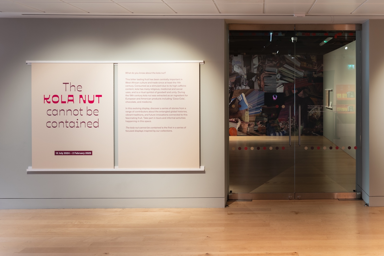 Photograph of a large panel outside a gallery to the left of a glass entrance door with 'The Kola Nut Cannot be Contained' title in view.