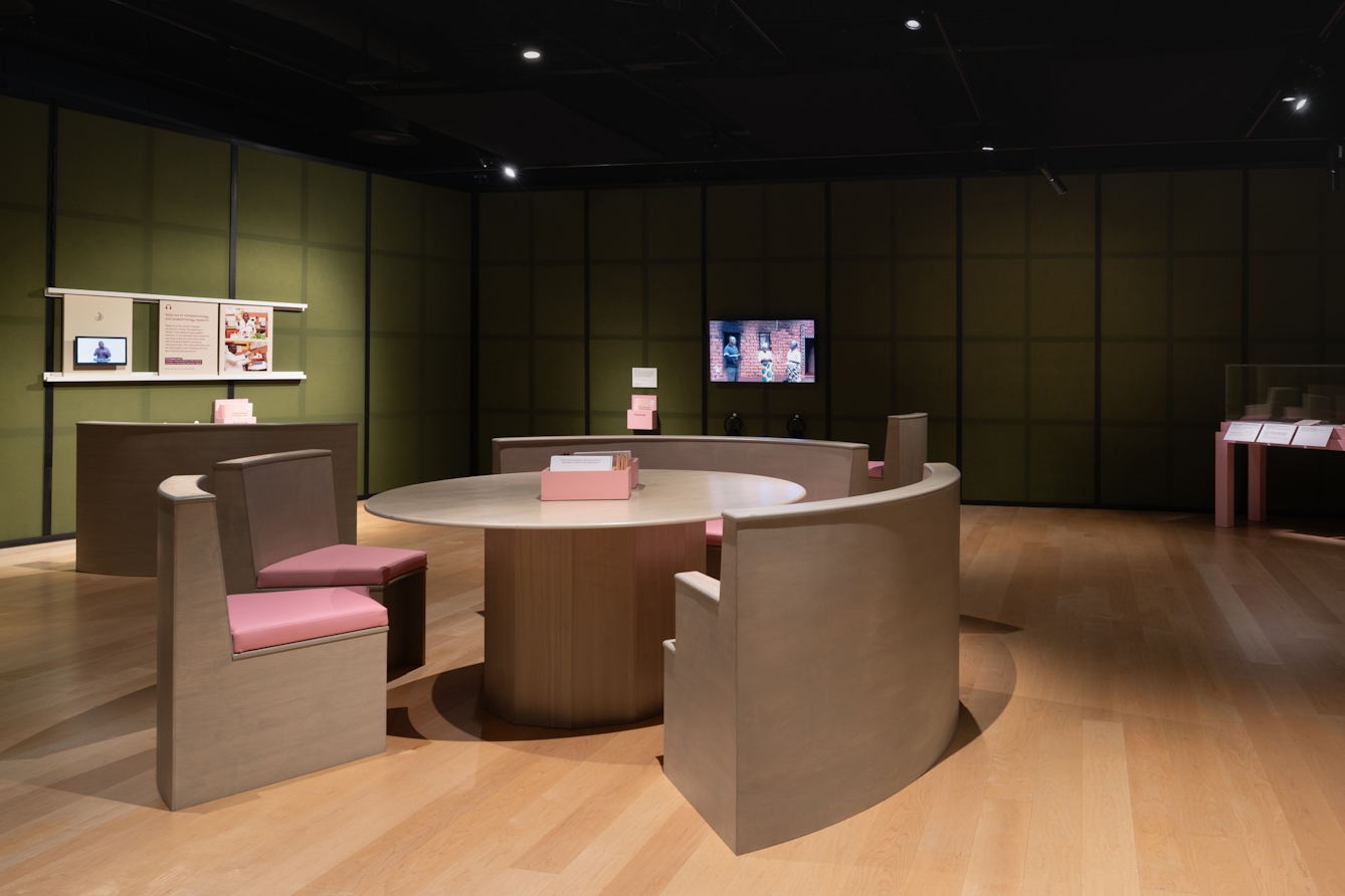 Photograph of an exhibition space featuring a large round table and chairs in the centre of the room; a BSL-interpreted video for an audio piece and another video on the adjacent wall.