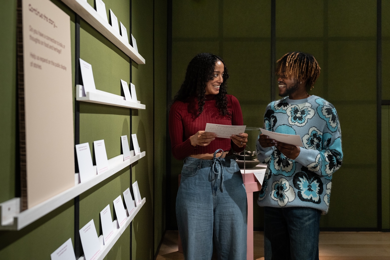 Photograph of two people talking and reading postcards from a shelf on the wall of a gallery space.