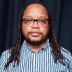 Head and shoulders photograph of a man with long hair, glasses and facial hair looking to camera.