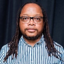 Head and shoulders photograph of a man with long hair, glasses and facial hair looking to camera.