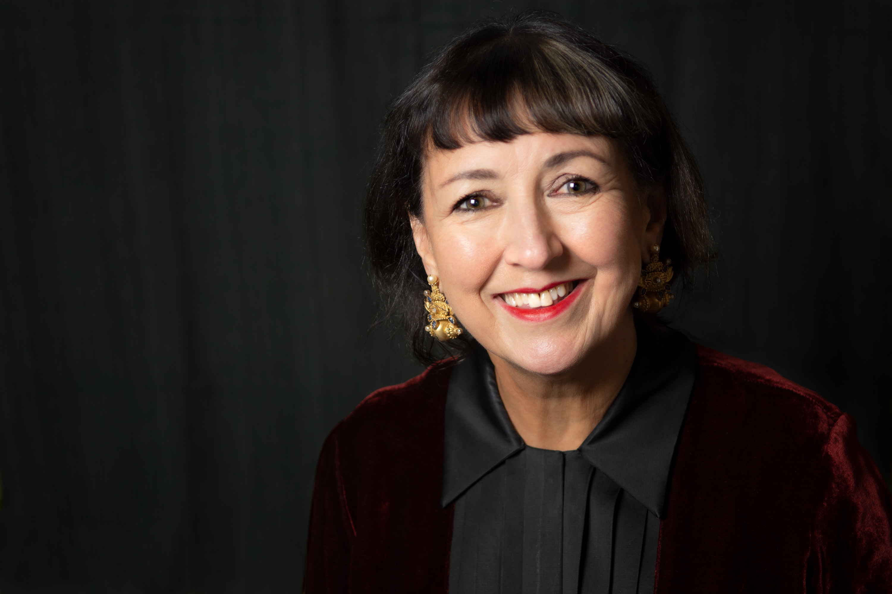 Head and shoulders photo of a woman with shoulder length brown hair wearing a red velvet jacket and black blouse