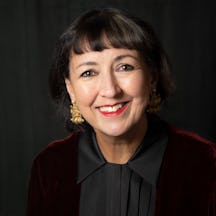 Head and shoulders photo of a woman with shoulder length brown hair wearing a red velvet jacket and black blouse