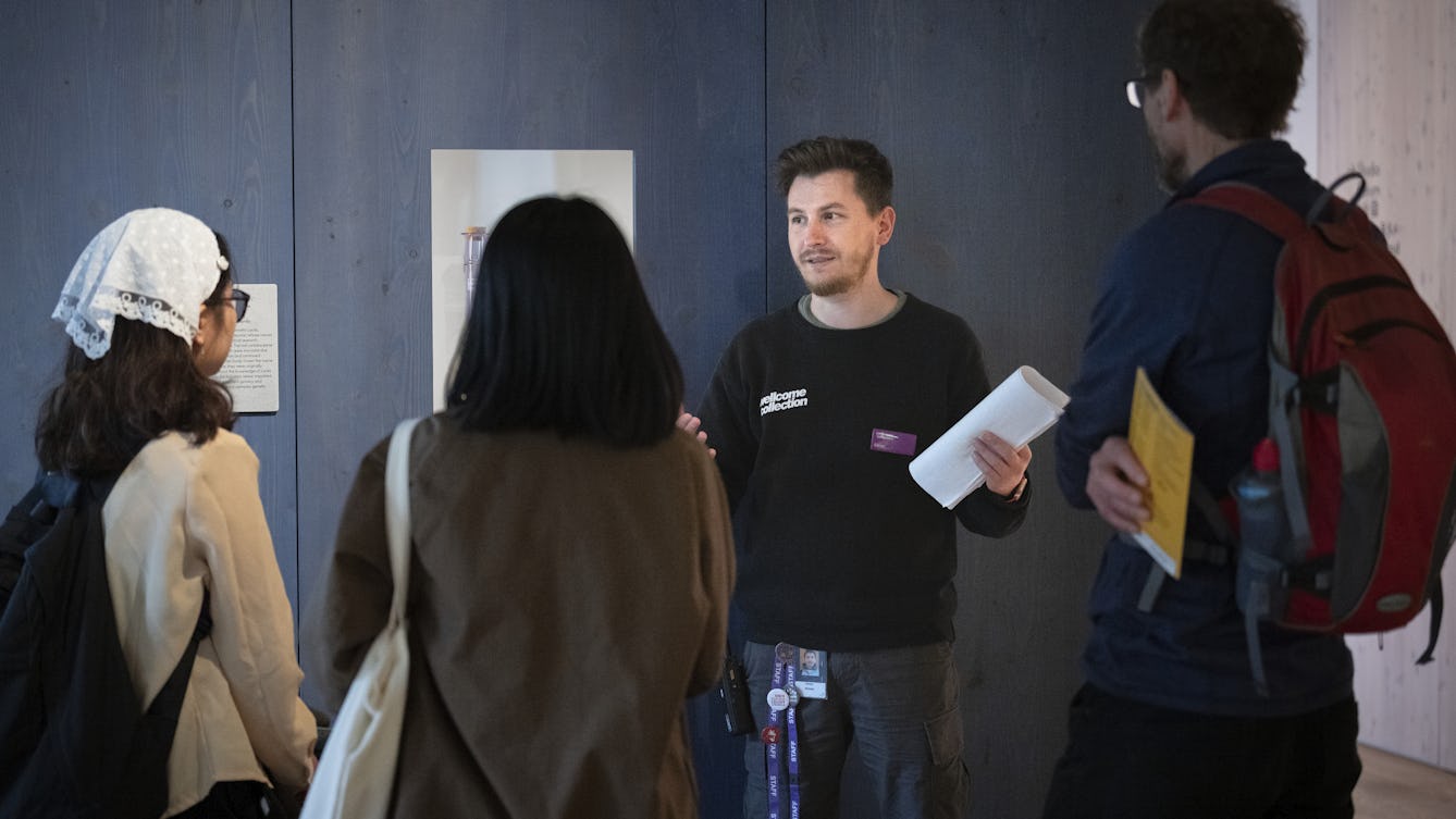 A visitor experience assistant wearing a black T-shirt with 'Wellcome Collection' written on it, giving a tour of the exhibition space. They are stood in front of a wall displaying framed exhibits, their hands raised in explanation. In front of them are members of the public listening to them speak.