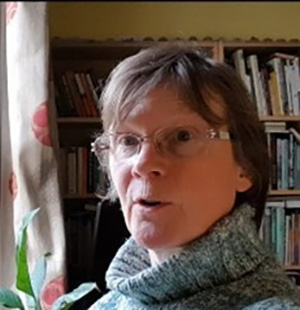 Black and white photo of the head and shoulders of an older woman with short grey hair and glasses. she is looking towards the camera and talking.