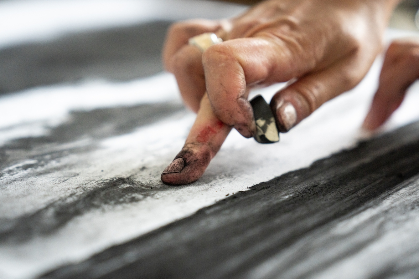 A close-up photograph of hand, dirty with charcoal dust with an extended finger rubbing across the surface of a charcoal artwork.