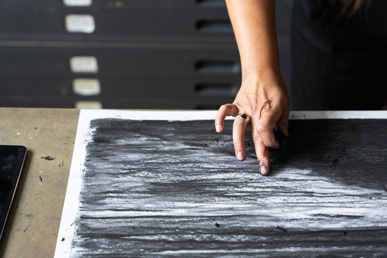 A close-up photograph of hand, dirty with charcoal dust with an extended finger rubbing across the surface of a large scale charcoal artwork.