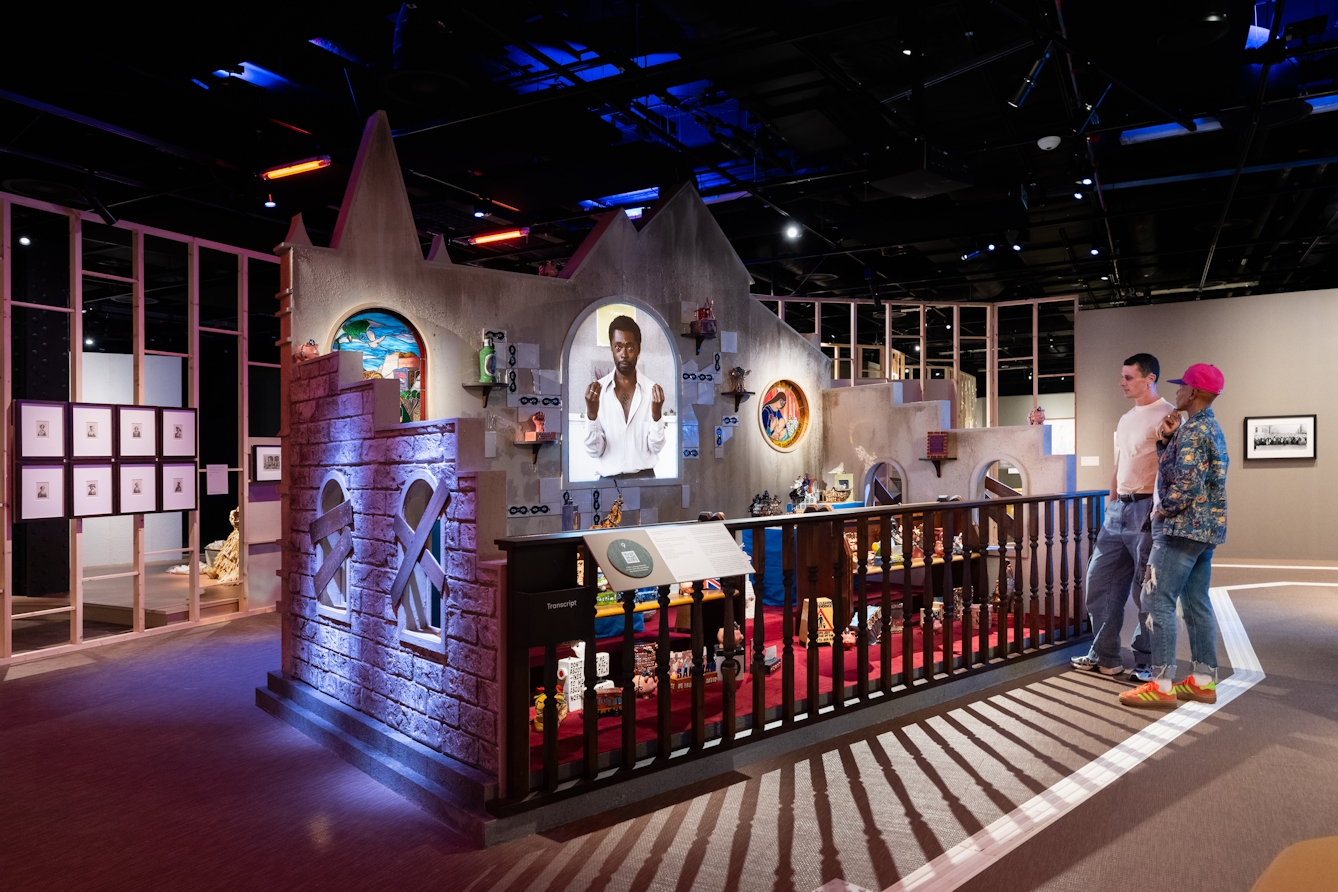 A photograph of two people in an exhibition space looking at a large scale installation artwork. The artwork shows the inside of a church with stained glass windows and a film  being projected in the centre. There are also wooden pews with ceramic works on display. 