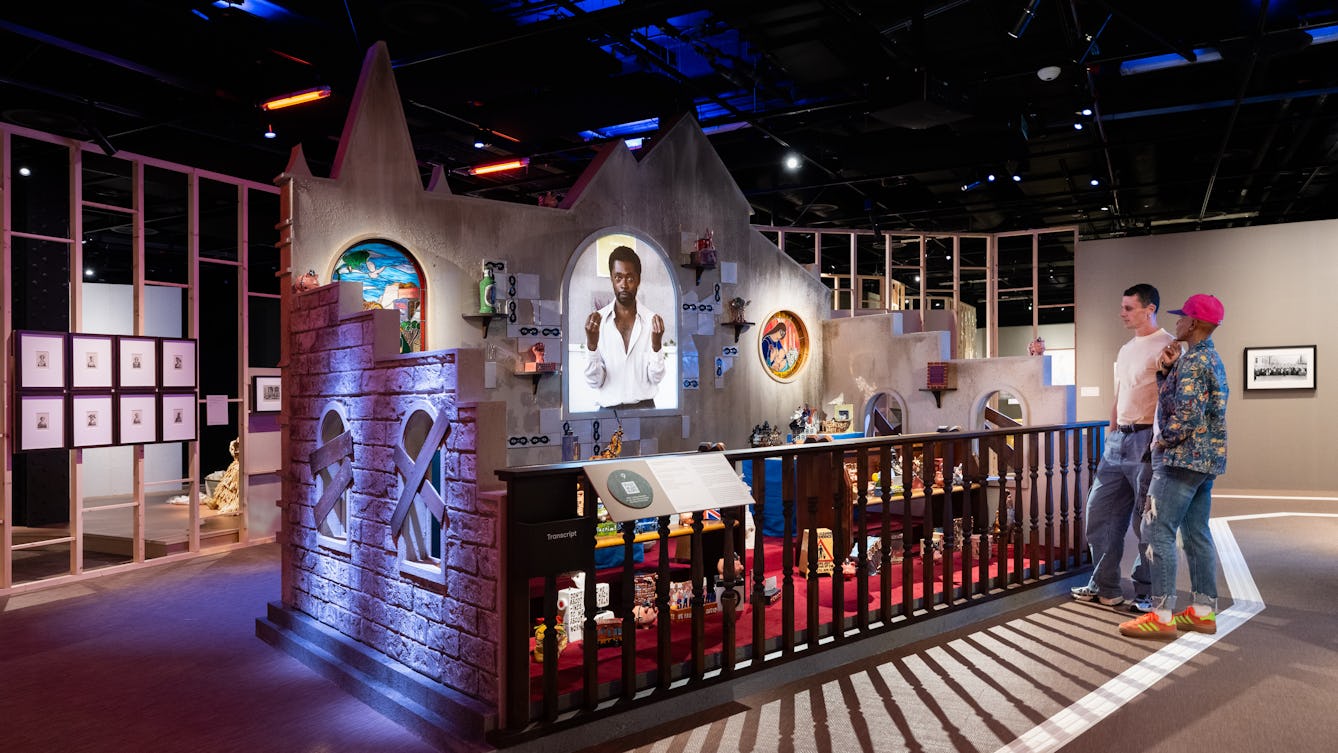 A photograph of two people in an exhibition space looking at a large scale installation artwork. The artwork shows the inside of a church with stained glass windows and a film being projected in the centre. There are also wooden pews with ceramic works on display.