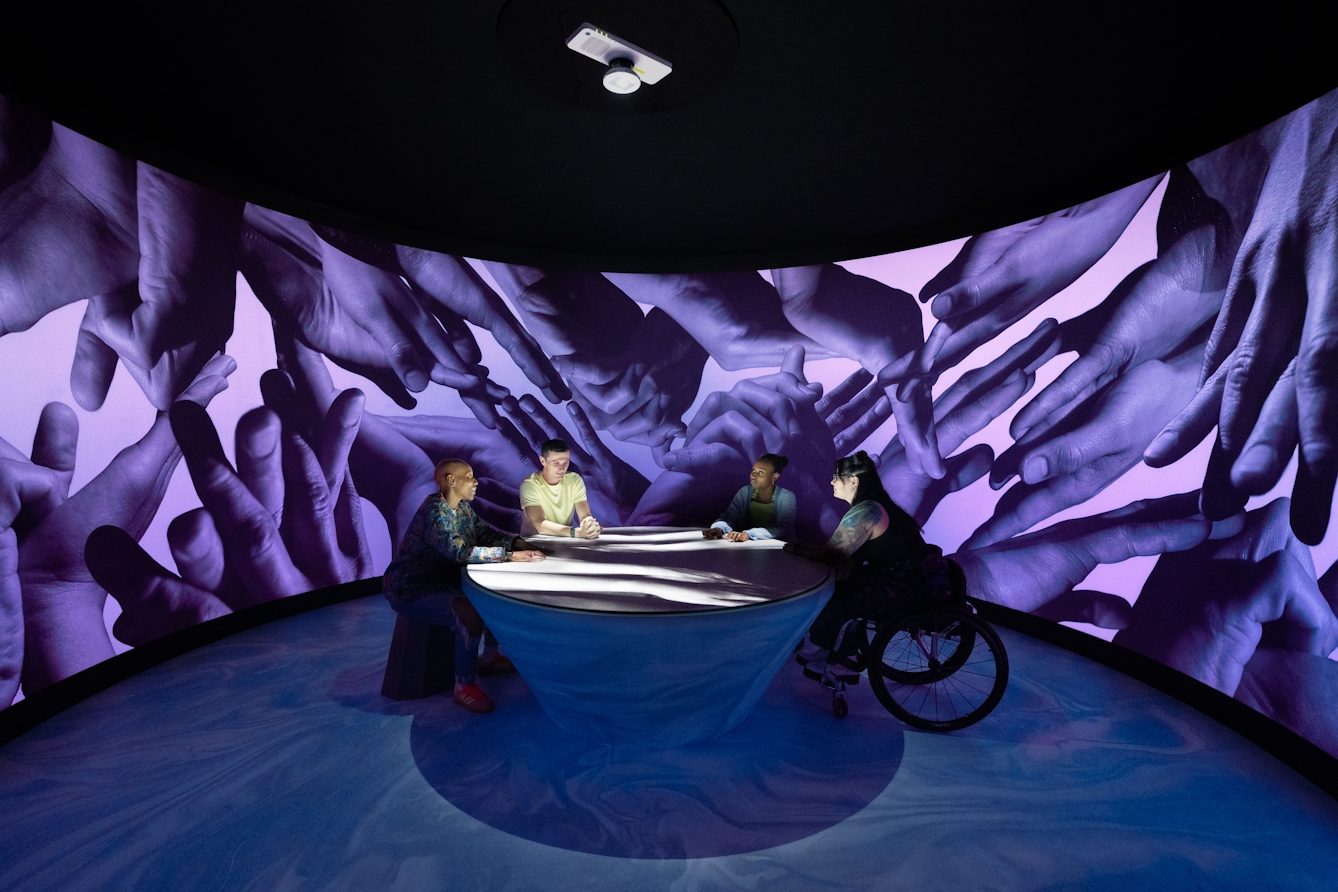 A photograph of four people sitting around a table within a large scale art installation. There is a black and white film being projected on to the table and across their hands. They are surrounded by a curved wall covered with a textile wallpaper depicting photographic representations of hands, lit by purple LEDs.