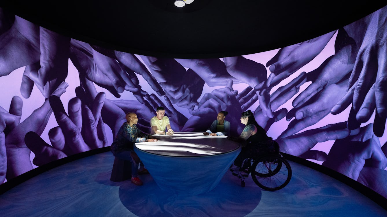 A photograph of four people sitting around a table within a large scale art installation. There is a black and white film being projected on to the table and across their hands. They are surrounded by a curved wall covered with a textile wallpaper depicting photographic representations of hands, lit by purple LEDs.