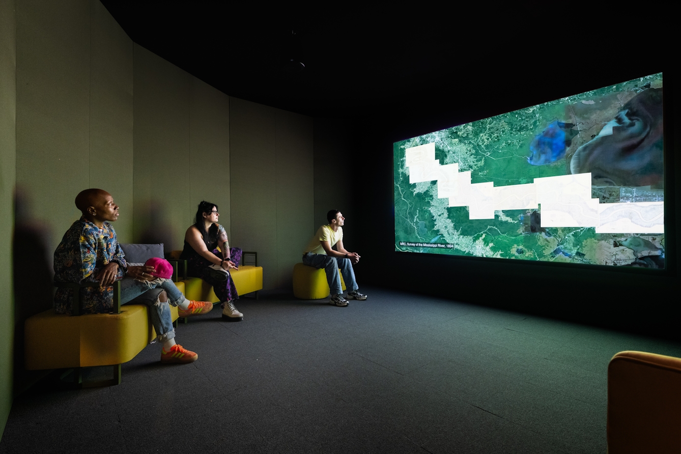 A photograph of three people sitting on chairs looking at a large film projection. The film show a satellite map with overlayed graphical information regarding pollution.