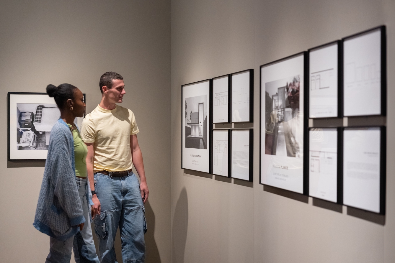 A photograph of two people looking at a series of framed artworks. The works are black and white photographs of buildings accompanied by architectural plans.