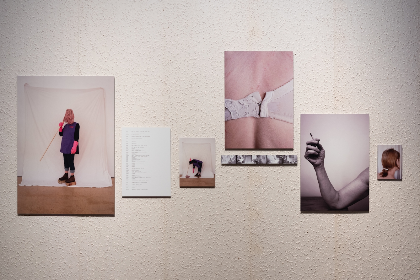 A photograph of a display of colour photographic prints, they are arranged in clusters and depict staged portraits of domestic cleaners and abstract images of the body. They are displayed against a wall of aged textured wallpaper.