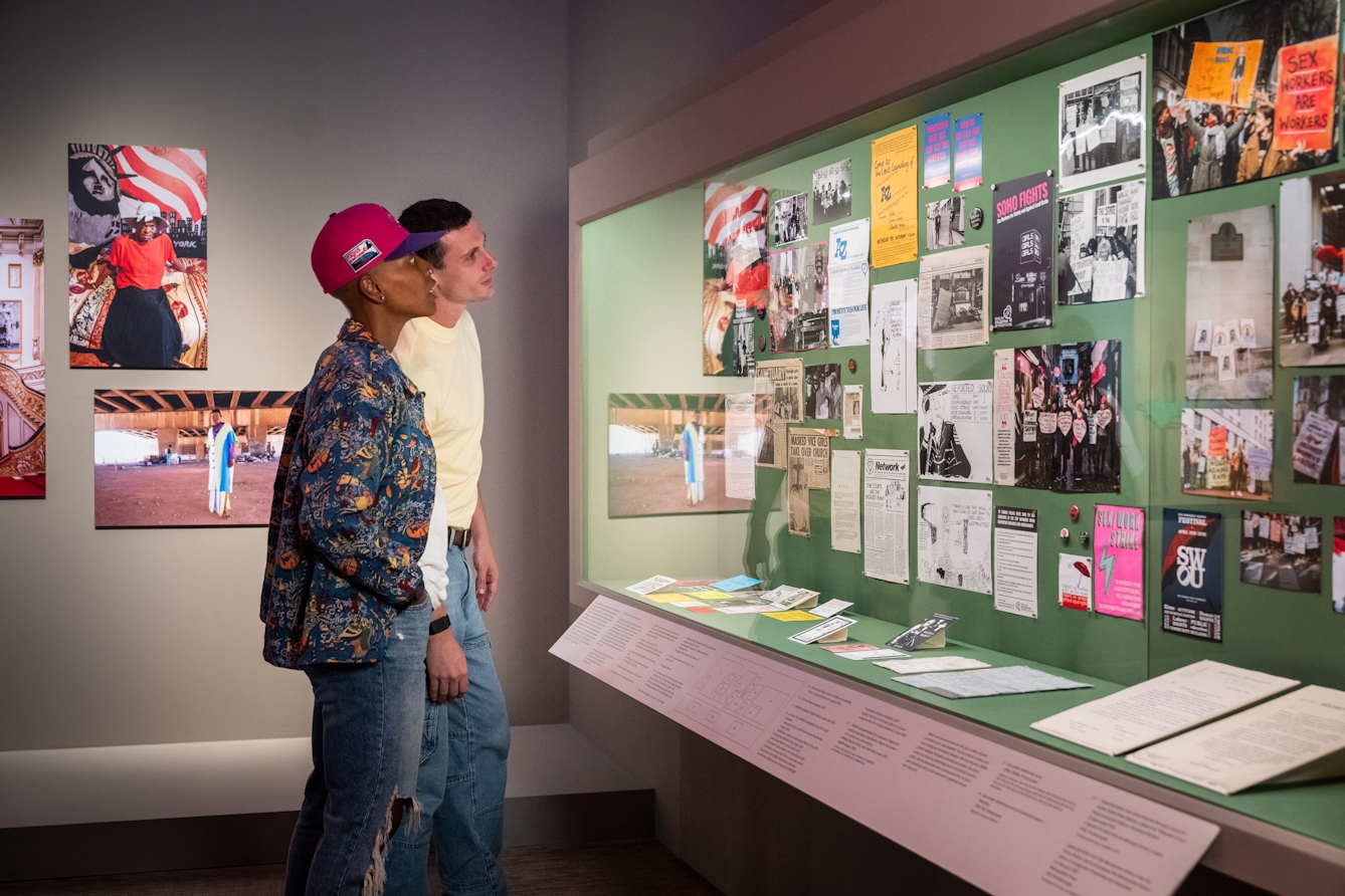 A photograph of two people looking at a display case of archive material which includes posters, photographs and leaflets relating to sex work. In the background are large photographic prints.