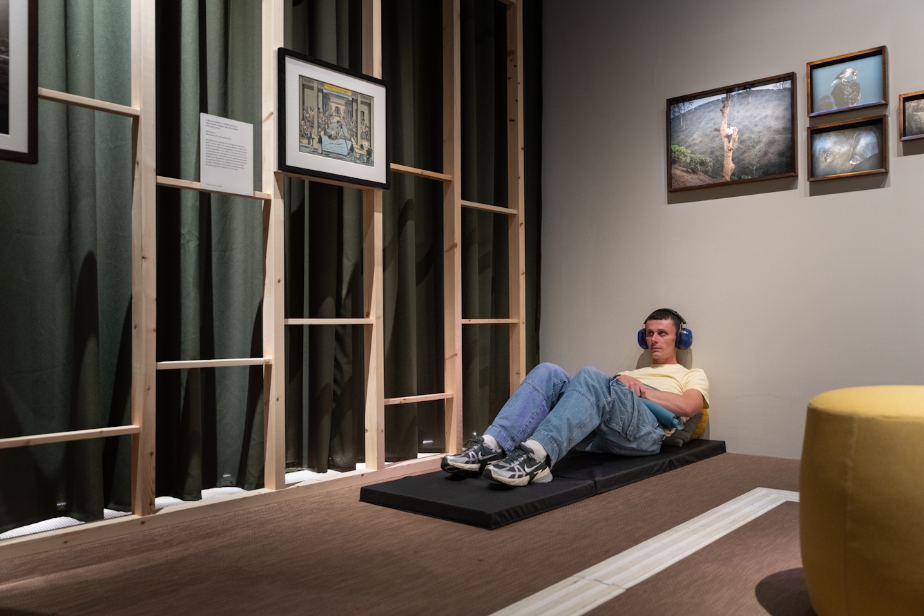 Photo of a person at a relaxed opening of the exhibition 'Hard Graft: Work, Health and Rights'. They are wearing ear defenders and sitting on a comfortable mat in the exhibition space.