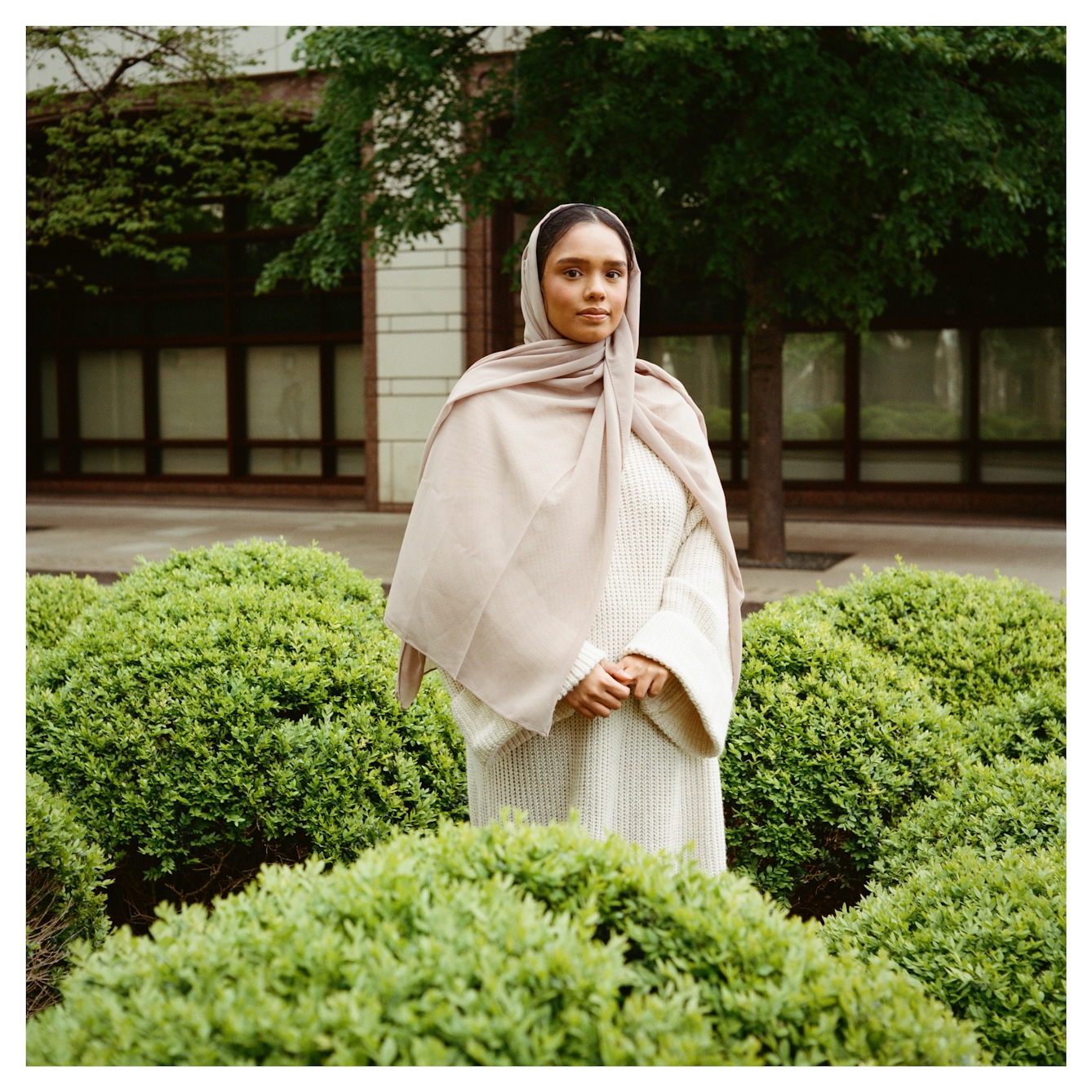 Portrait photograph of Atifa, a woman of Bengali Muslim heritage standing outside surrounded by low lying bushes. Her hands are loosely clasped together across her body as she gazes straight to camera with a warm, open expression.