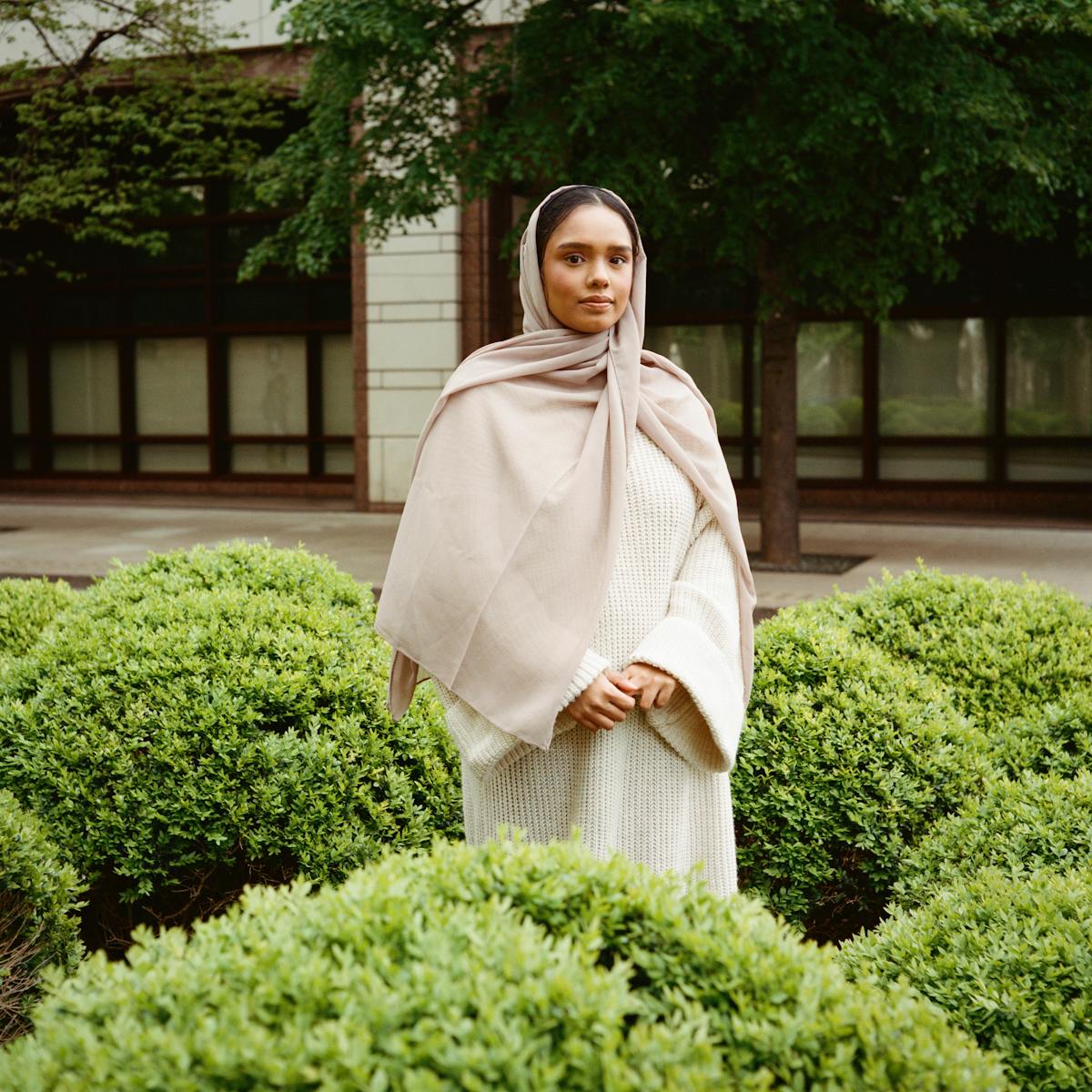 Portrait photograph of Atifa, a woman of Bengali Muslim heritage standing outside surrounded by low lying bushes. Her hands are loosely clasped together across her body as she gazes straight to camera with a warm, open expression.