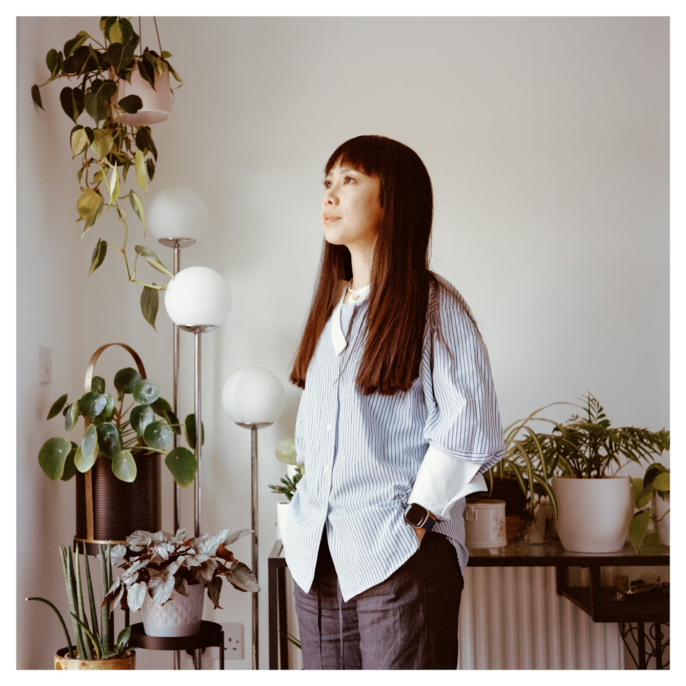 Portrait photograph of Hazel, a woman of Chinese heritage standing in the living room of her home. Her hands are buried in her trouser pockets and she is surrounded by pots of house plants. She gazes into the distance off to the left.