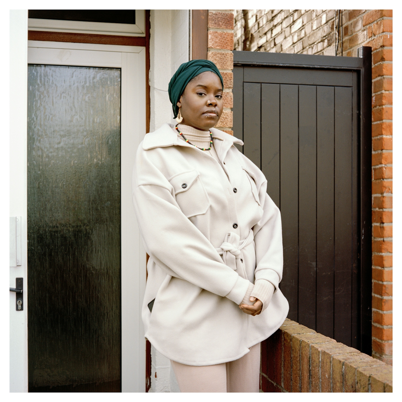 Portrait photograph of Marsha, a Black woman of Afro-Caribbean heritage standing outside the front door of her home. Her hands are clasped front of her and she is looking straight into camera with a strong, calm expression.