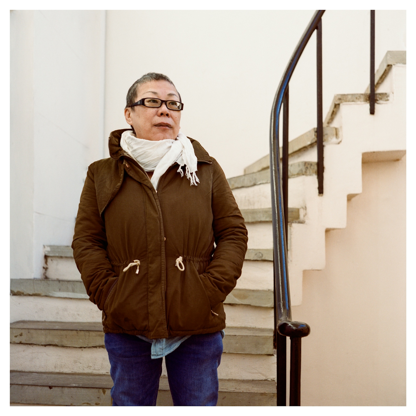 Portrait photograph of Jia-Ai, a woman of Chinese heritage standing outside at the base of a curved staircase. She is dressed in a winder coat and scarf, her hands buried the jacket pockets. She gazes into the distance off to the right.