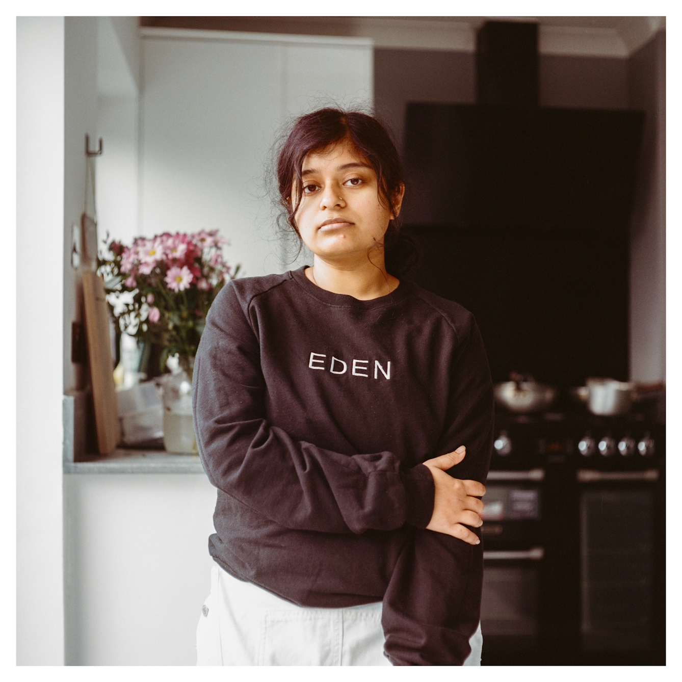 Portrait photograph of Tasmiyah, a woman of Bangladeshi/Bengali heritage standing in a kitchen. Her arm is held across her torso and the word 'Eden' is written in white on her black jumper. She is looking straight into camera with a natural expression.