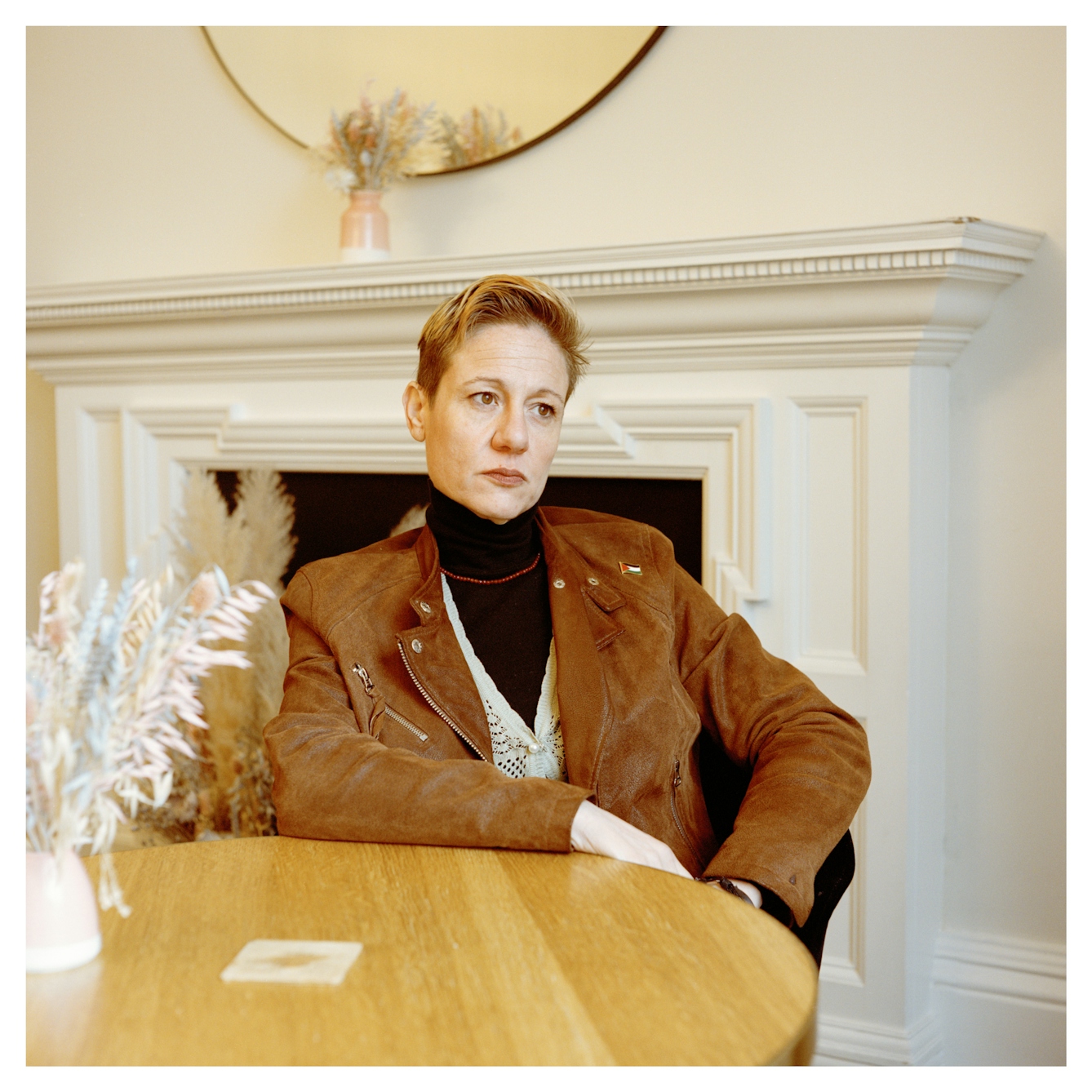 Portrait photograph of Suzan, a woman of Turkish Cypriot British heritage sitting at a table in front of a large mantlepiece. She leans on the table with one arm, gazing into the distance off to the right.