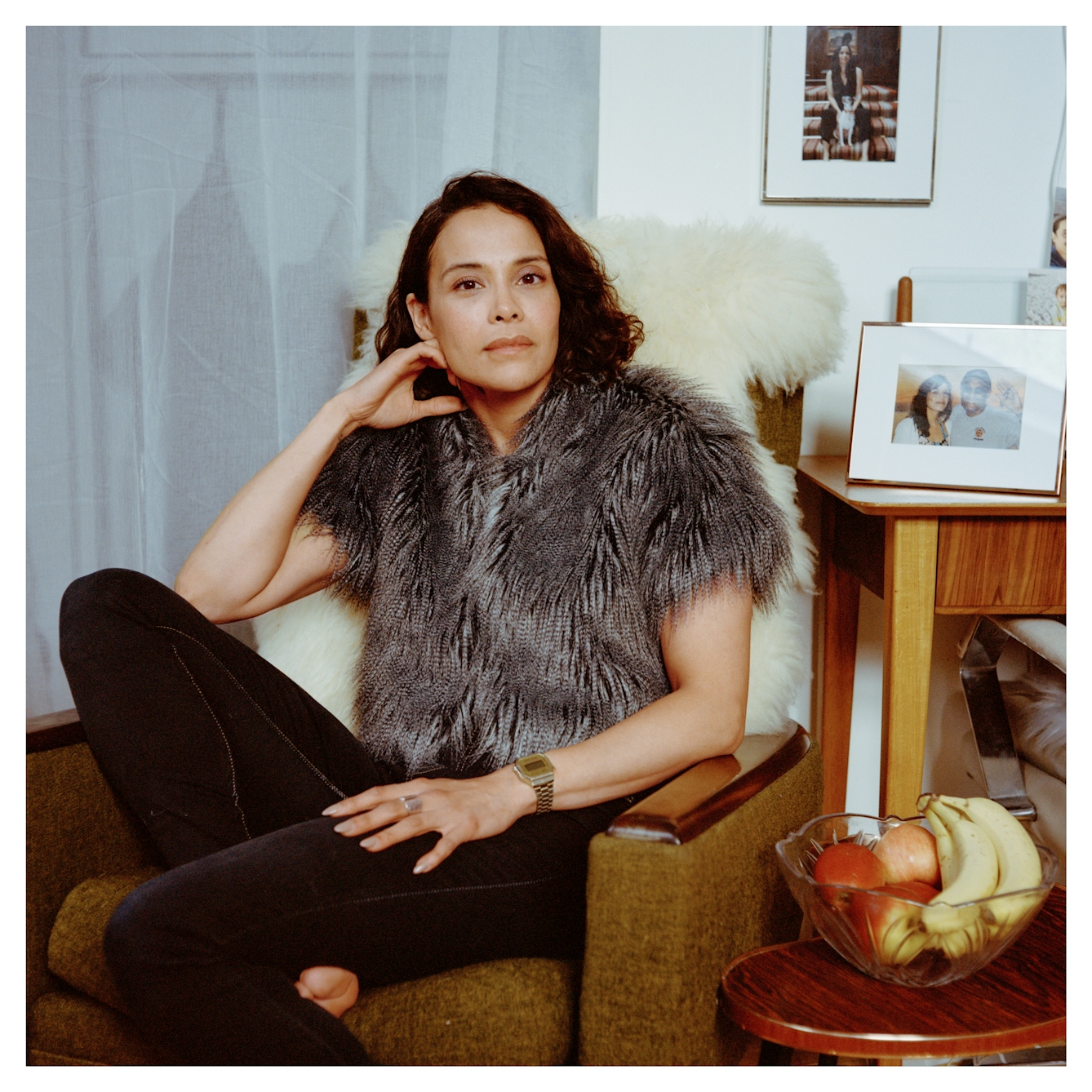 Portrait photograph of Valerie, a woman of East Asian heritage sitting in an armchair next to a table of framed family photos. Her head is resting lightly on her hand as she looks straight into camera with a calm open expression.