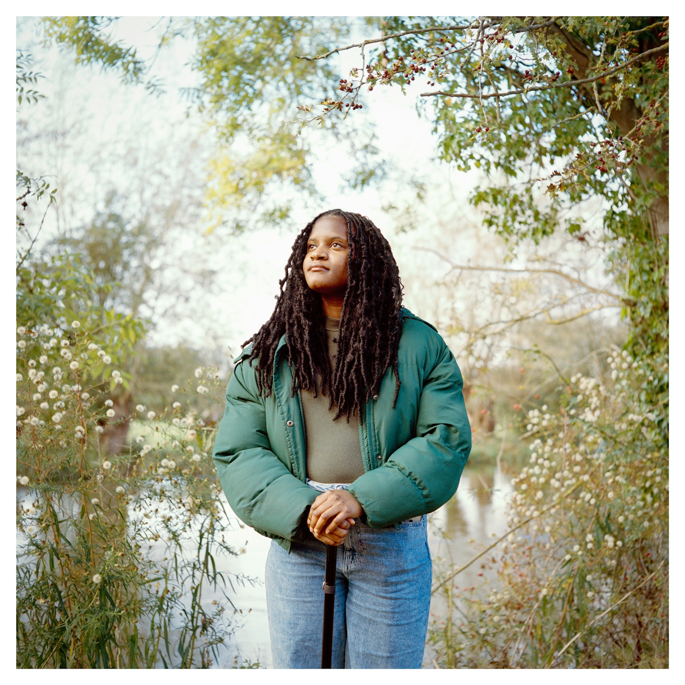 Portrait photograph of Savannah, a Black woman with long hair, standing in a parkland wearing a green puffer jacket, hands gently clasped on a stick. She is gazing off into the distance to the left.