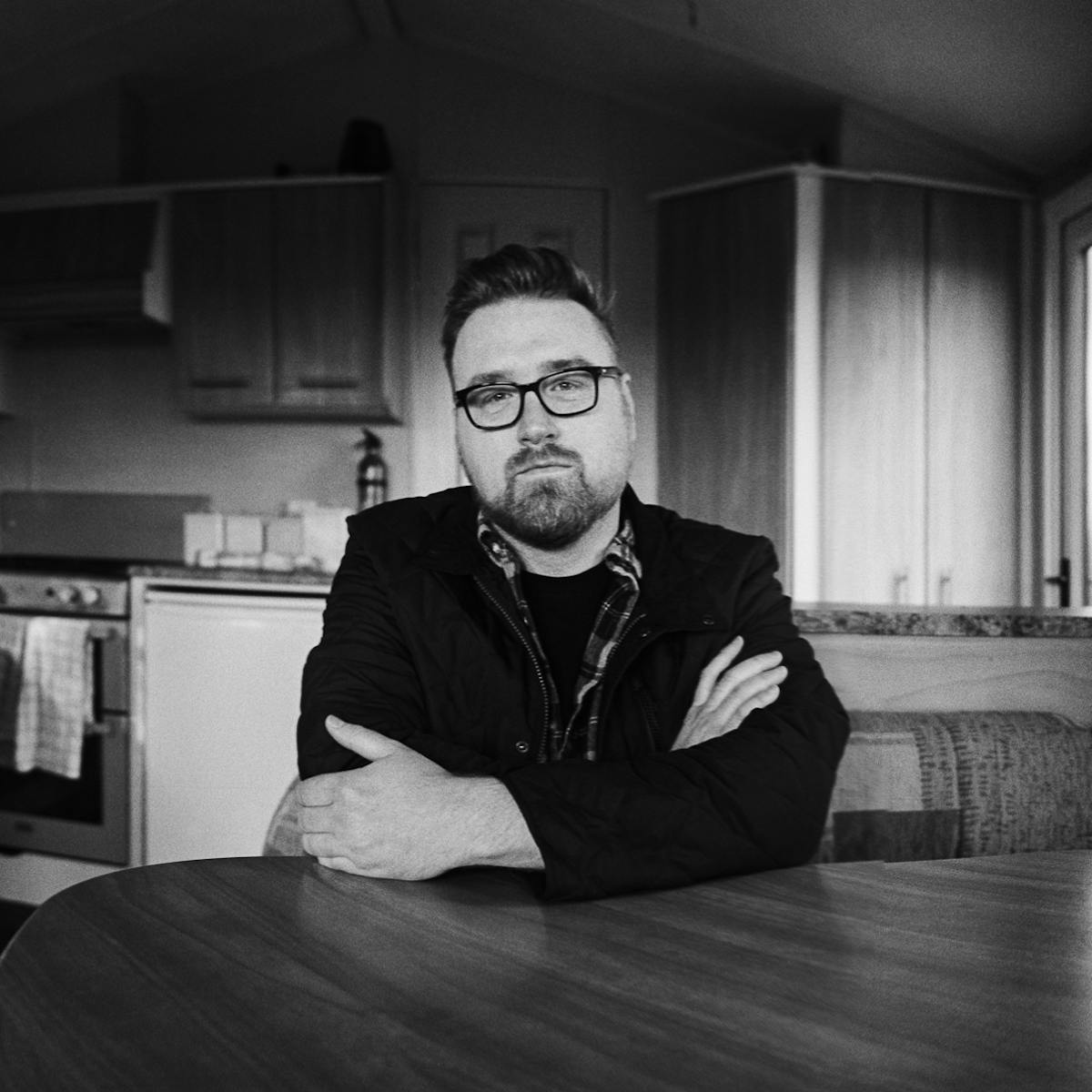 Black and white panoramic portrait of an individual sitting at their kitchen table, arms folded, looking straight to camera. Behind them are the kitchen appliances and countertops.
