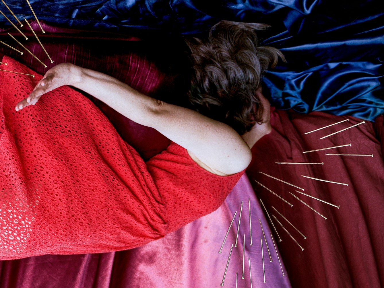 Detail from a larger artwork created with a colour photographic print of a female figure in a bright red dress, set against a purple and blue draped silk background. She is lying horizontally in the frame, face down, caught as if in mid fall.  Her hair and dress are blown upwards. Her body is surrounded by groups of dress pins, laid on top of the photographic print. The pins are arranged as if they are a flight of arrows directed at her body. One group attacks her neck and chest.