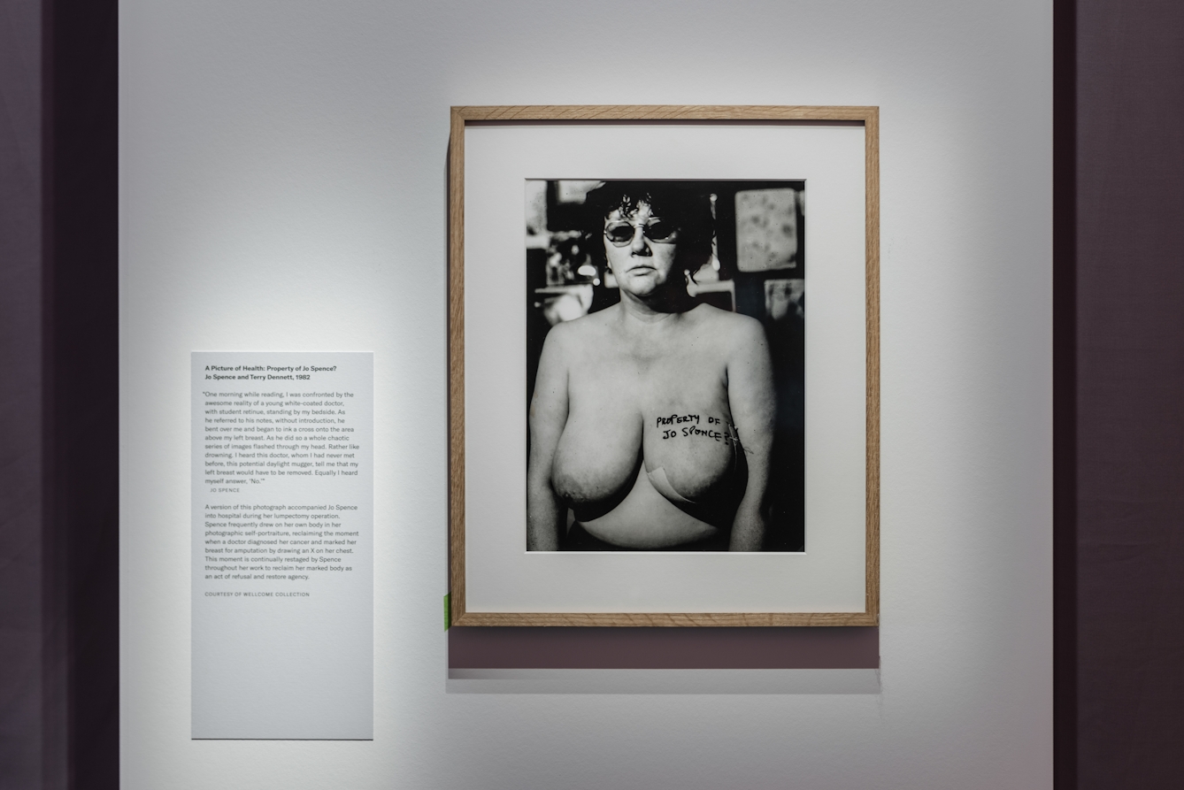 Photograph of a wall in a gallery showing a framed black and white photograph of a woman, naked from the waist up. On her left breast is written the words, 'Property of Jo Spence?' To the left of the photograph is a text panel, attached to the wall.