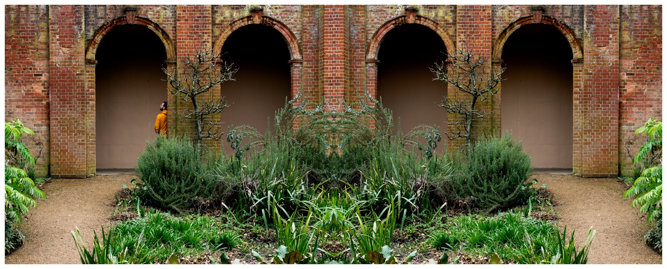 Photographic panorama showing an arched brick walkway and planted flowerbeds. The panorama is mirrored down its vertical centre line except that a small human figure in a yellow top leaning against the archway appears in the left half, but not in the right half.