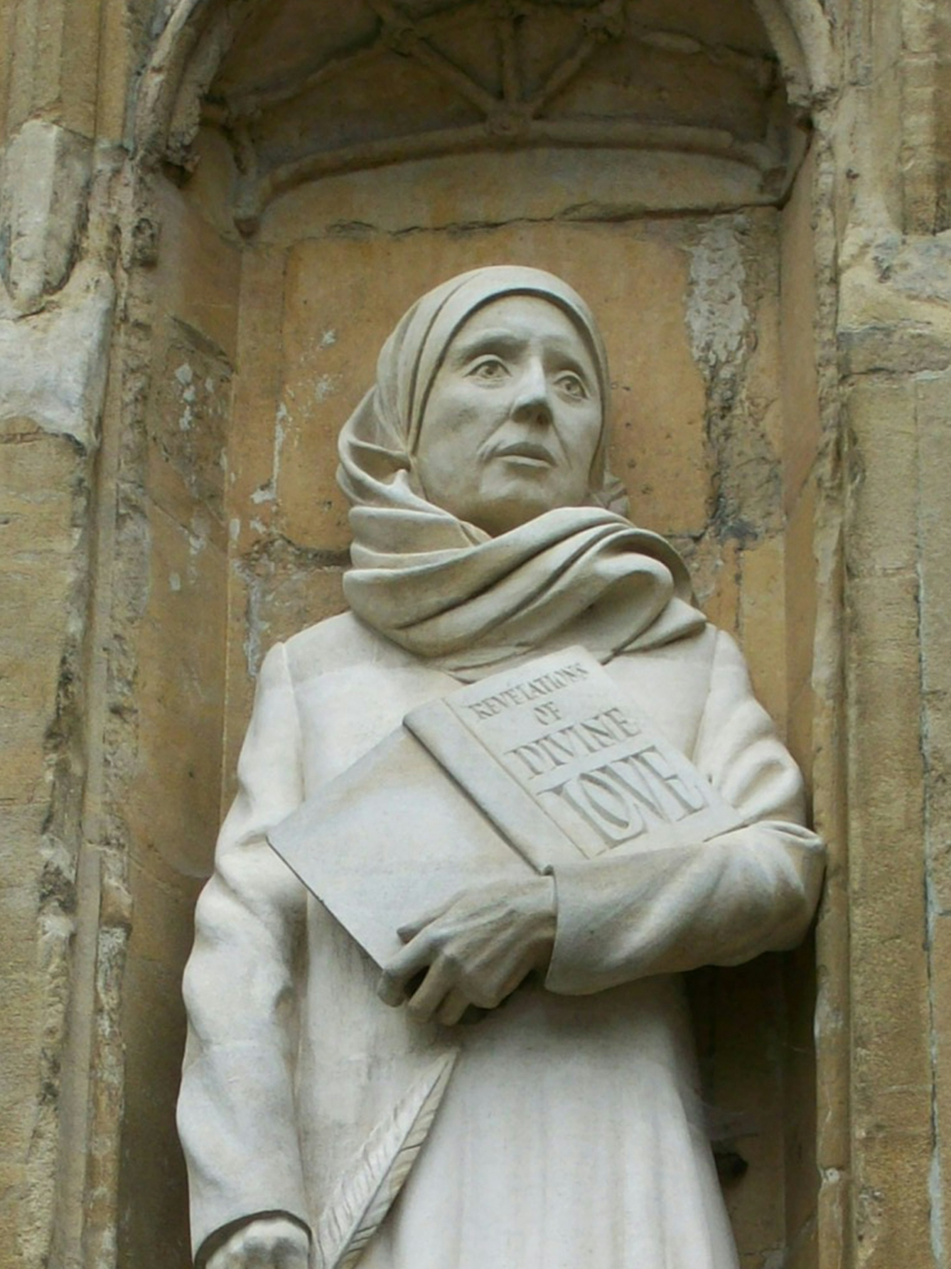 Photograph of white stone statue of Julian of Norwich. She is holding a book and the title reads 'Revelations of Divine Love'. The statue is ensconced in a stone alcove. 