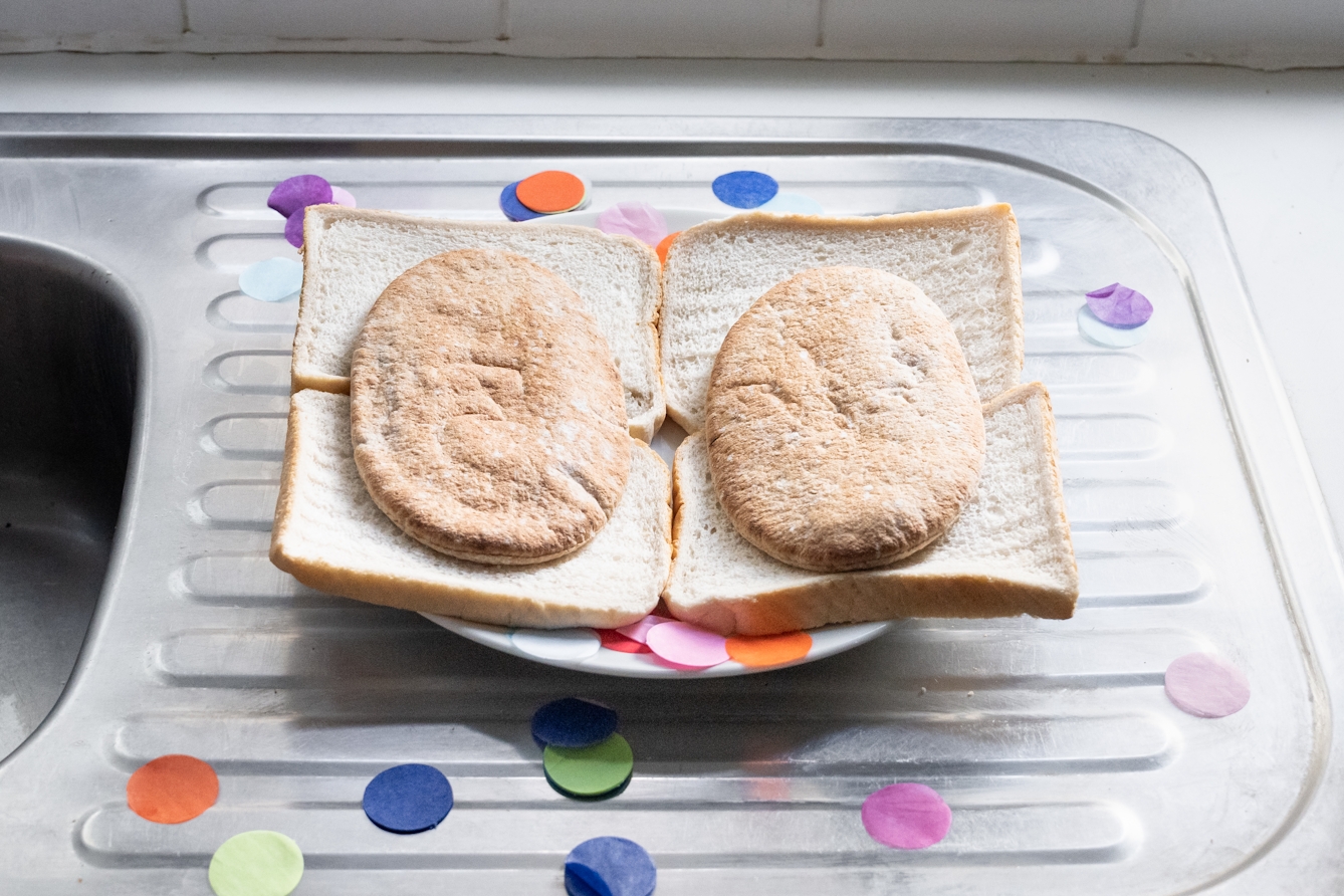 A photograph of a kitchen sink draining board, on which is placed a plate stacked with layers of sliced white bread and pitta bread.