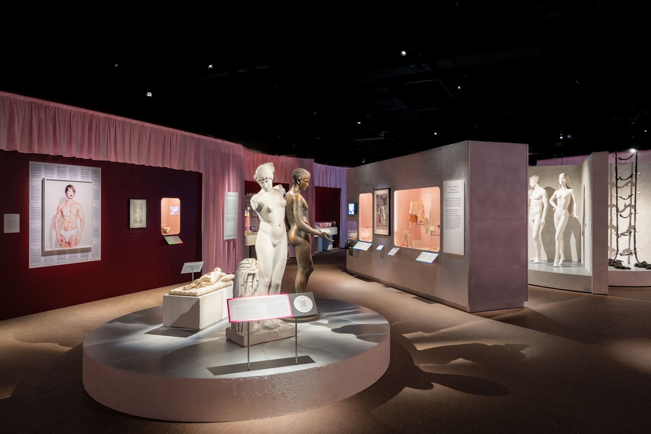 An area in an exhibition space showing images on walls along side objects displayed behind glass. In front of these is a circular dias with classical male and female figures, both standing and a reclining figure on a plinth.. 