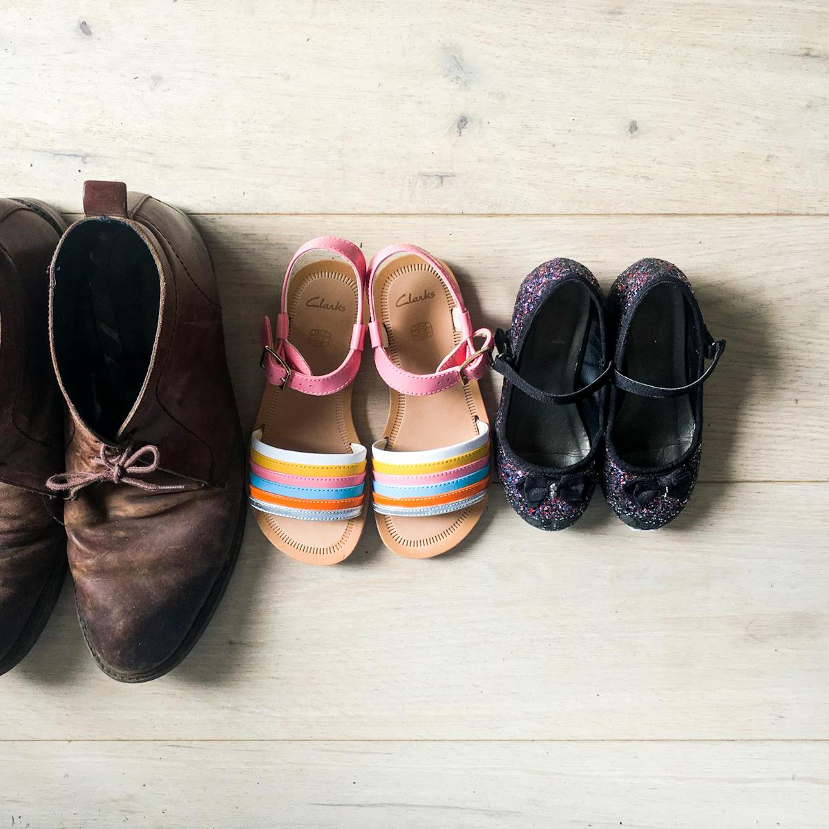 Photograph of a 'bird's eye' view of three pairs on shoes on a lime washed floor-boarded floor. The shoes are lined up in size order from largest on the left and smallest on the right. The largest are a pair of adults brown suede shoes with the laces tied and worn toe caps. In the middle are a pair of girl's sandals with rainbow coloured front straps and pink angle straps. On the far right are the smallest shoes, a pair of sparkly purple girls ballerina style pumps, with small black bows on the toes. 