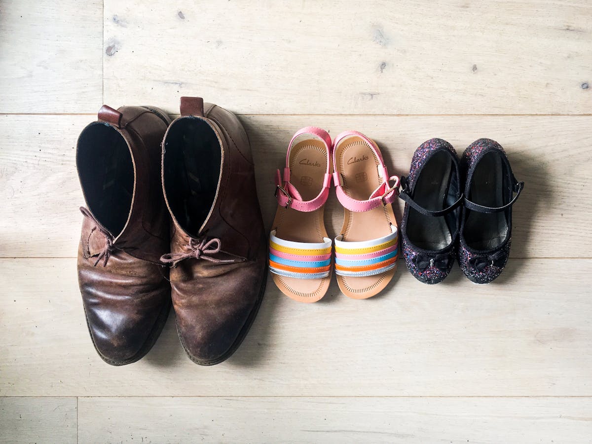 Photograph of a 'bird's eye' view of three pairs on shoes on a lime washed floor-boarded floor. The shoes are lined up in size order from largest on the left and smallest on the right. The largest are a pair of adults brown suede shoes with the laces tied and worn toe caps. In the middle are a pair of girl's sandals with rainbow coloured front straps and pink angle straps. On the far right are the smallest shoes, a pair of sparkly purple girls ballerina style pumps, with small black bows on the toes. 