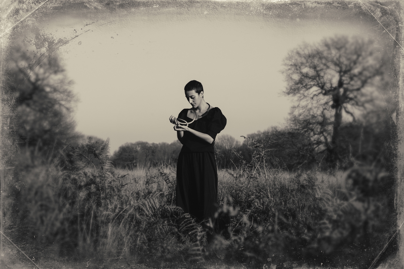 Sepia toned photograph with a digital filter applied to give the impression the photograph was created using a glass-plate process from the mid 19th century. The effect of this filter includes scratches and fingerprints. The photograph shows a woodland scene made up of tall grasses, bracken and distant trees. In the centre of the image stands a tall woman dressed in a black floor length dress. She is looking down at her hands which are raised in front of her chest. Wrapped around her neck and running over her shoulder and down to her open hands is a long darkly toned snake. The centre of the frame is in sharp focus, but the edges descend quickly into a blur.