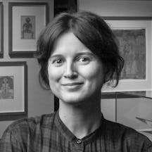 Black and white photographic head and shoulders portrait of Bárbara Rodríguez Muñoz standing in an exhibition gallery space. She is wearing a checked dress with her arms folded across her front. Behind her are framed artworks on the walls.